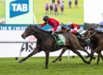 A dark brown racehorse with a red-jacketed jockey leads a crowded horse race on a grassy track, TAB signage in the background.