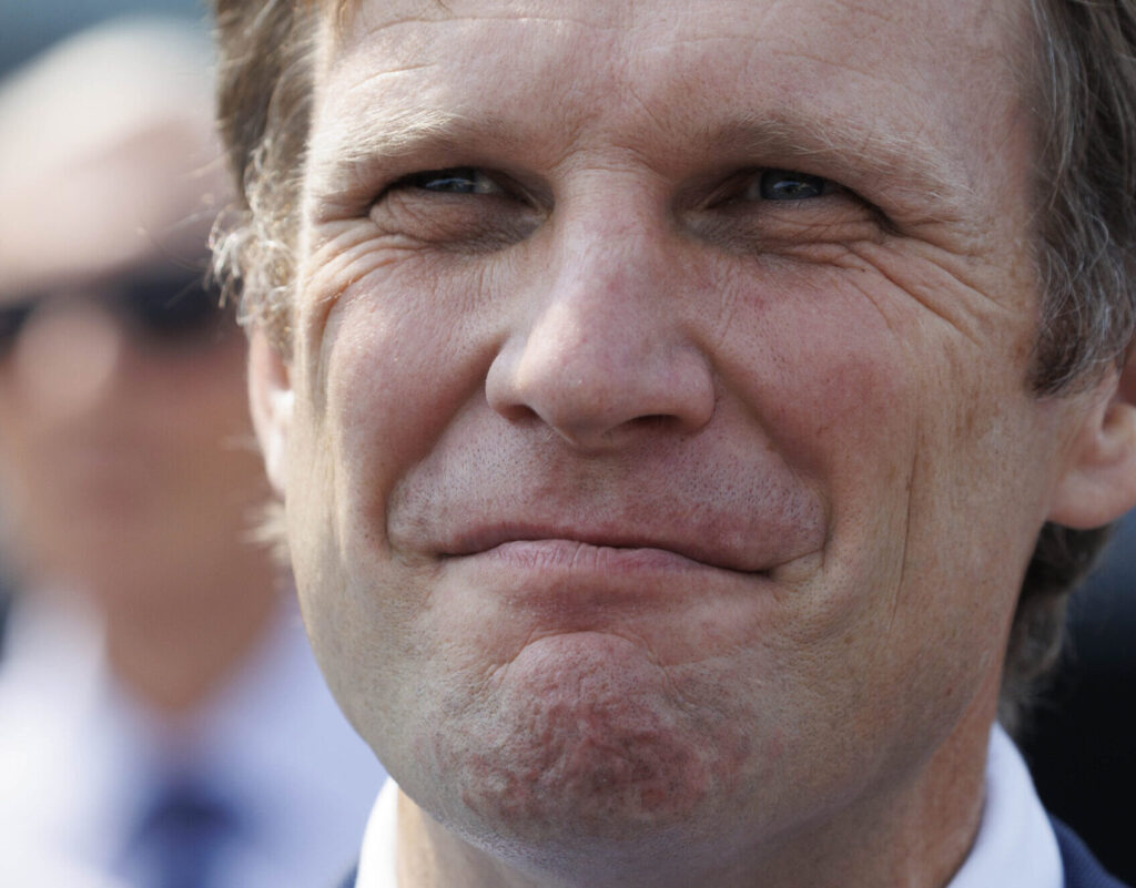 Close-up of a middle-aged man's smiling face with light stubble and pronounced wrinkles around the eyes and mouth.