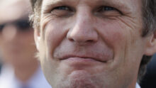 Close-up of a middle-aged man's smiling face with light stubble and pronounced wrinkles around the eyes and mouth.