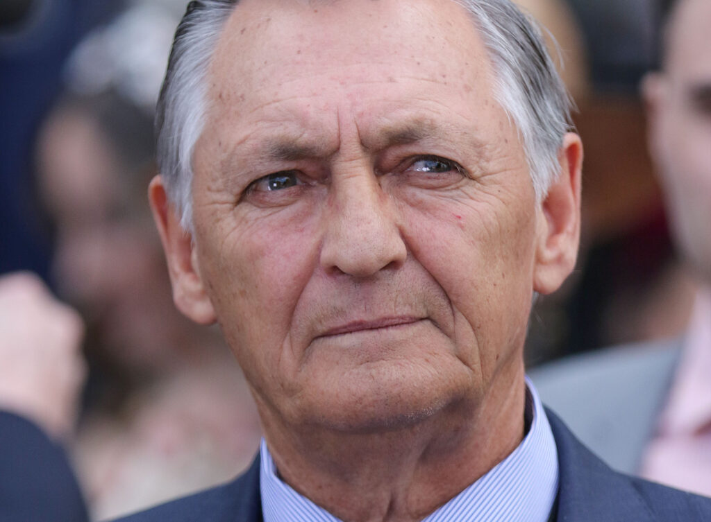Close-up of an elderly man's face in a dark suit and light blue striped shirt, looking slightly to the left.