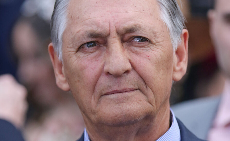 Close-up of an elderly man's face in a dark suit and light blue striped shirt, looking slightly to the left.