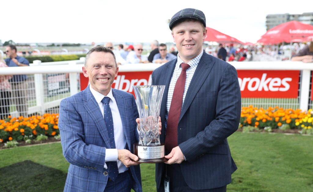 Two men in suits smile while holding a tall crystal trophy at a racecourse, with orange flowers and spectators in the background.