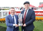 Two men in suits smile while holding a tall crystal trophy at a racecourse, with orange flowers and spectators in the background.