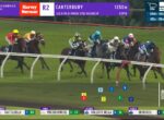 Thoroughbred horse race on a green turf track; jockeys in colorful silks push toward the right, with on-screen overlays showing race numbers, positions, and the current leader beneath a purple sponsor banner.