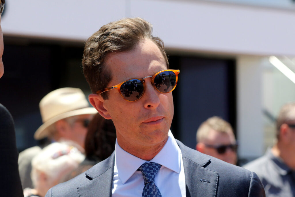 Man in a dark suit and blue patterned tie wears orange sunglasses at an outdoor event with blurred crowd in background, looking to the side.