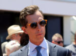 Man in a dark suit and blue patterned tie wears orange sunglasses at an outdoor event with blurred crowd in background, looking to the side.