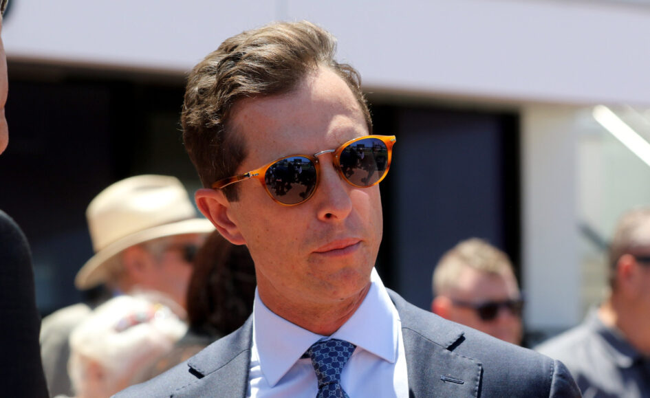 Man in a dark suit and blue patterned tie wears orange sunglasses at an outdoor event with blurred crowd in background, looking to the side.