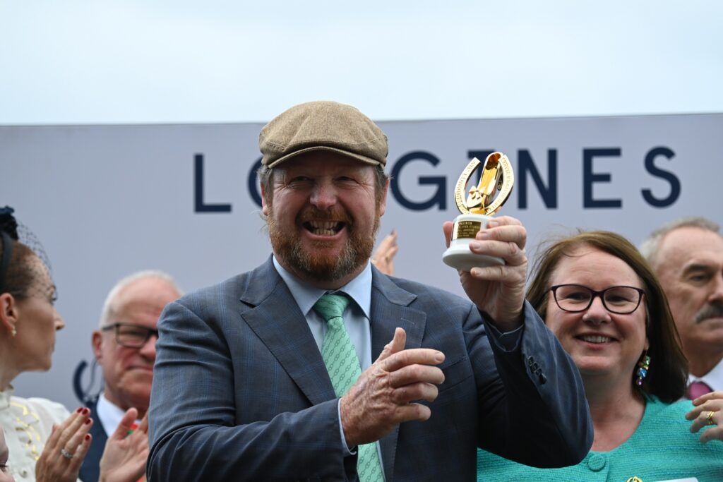 Smiling man in a gray suit and flat cap holding a gold trophy among a cheering crowd at an outdoor event.