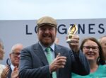 Smiling man in a gray suit and flat cap holding a gold trophy among a cheering crowd at an outdoor event.