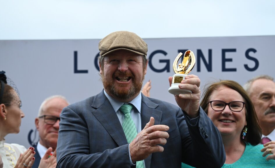 Smiling man in a gray suit and flat cap holding a gold trophy among a cheering crowd at an outdoor event.