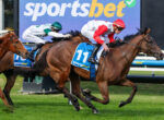 Three jockeys race on galloping horses on a grassy track, wearing green, red, and white silks with blue bibs and a stadium banner in the background.