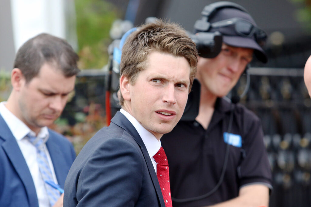Man in a navy suit and red tie glances at the camera at an outdoor event, with two colleagues in the background wearing headsets.