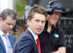 Man in a navy suit and red tie glances at the camera at an outdoor event, with two colleagues in the background wearing headsets.