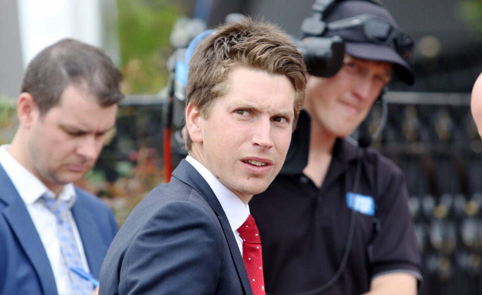 Man in a navy suit and red tie glances at the camera at an outdoor event, with two colleagues in the background wearing headsets.