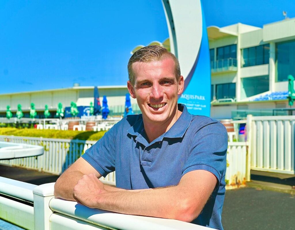 Man in a blue polo leaning on a white railing, smiling at the camera with a bright blue sky and water-park buildings in the background.