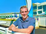 Man in a blue polo leaning on a white railing, smiling at the camera with a bright blue sky and water-park buildings in the background.