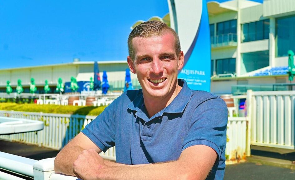 Man in a blue polo leaning on a white railing, smiling at the camera with a bright blue sky and water-park buildings in the background.
