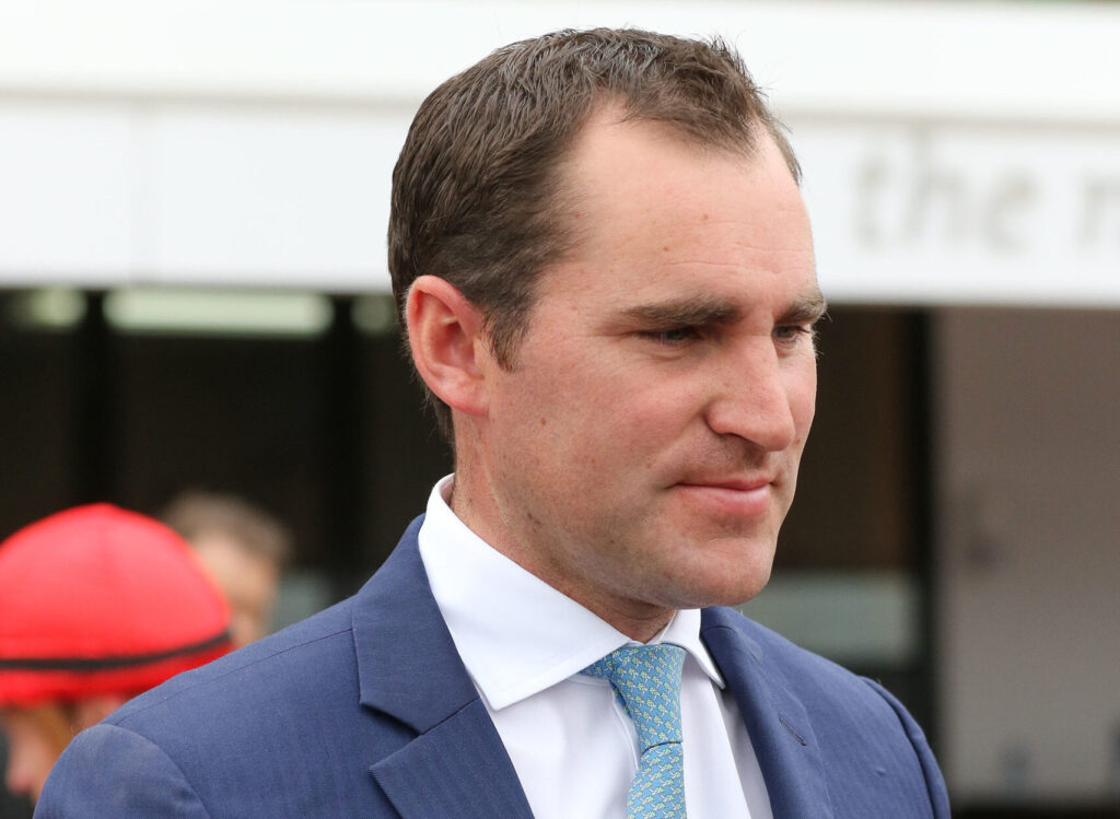 Man in a navy suit and light blue patterned tie, looking to the right at an outdoor event.