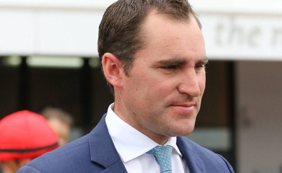 Man in a navy suit and light blue patterned tie, looking to the right at an outdoor event.