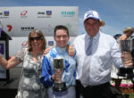 Three people celebrate on a stage, each holding a silver trophy and smiling at the camera, with sponsor banners in the background.