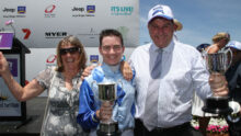 Three people celebrate on a stage, each holding a silver trophy and smiling at the camera, with sponsor banners in the background.