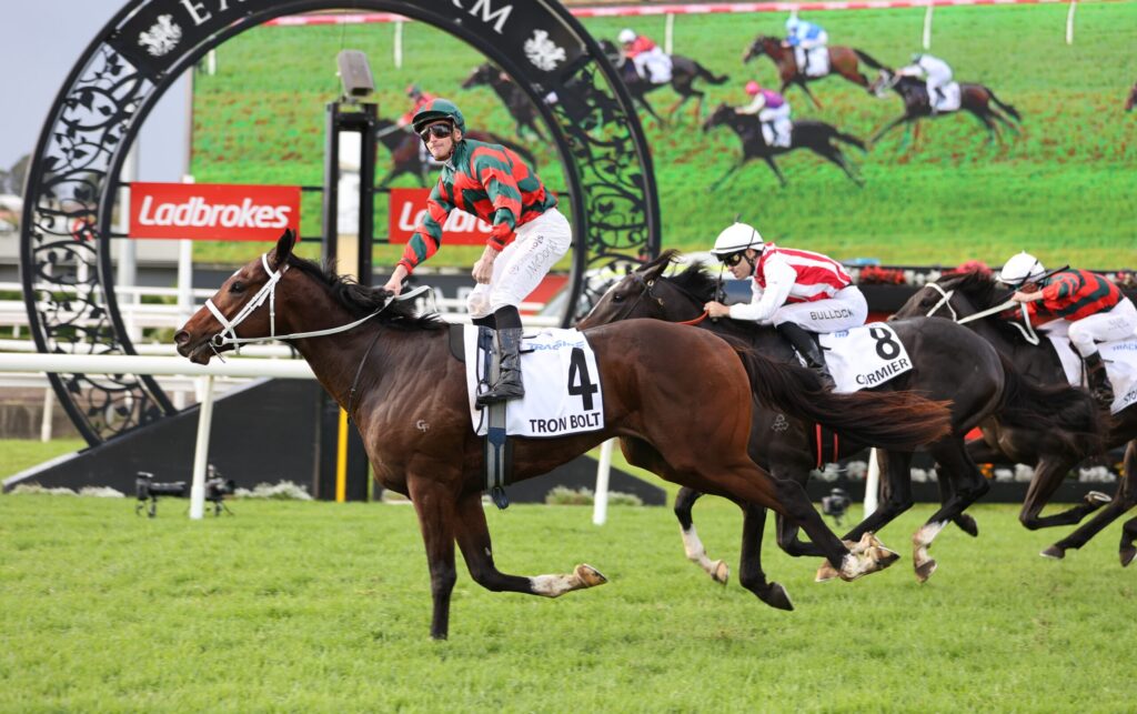 Jockey in red-green silks rides a brown horse with bib 4 ('Tron Bolt') leading a horse race on a green turf track.