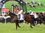 Jockey in red-green silks rides a brown horse with bib 4 ('Tron Bolt') leading a horse race on a green turf track.