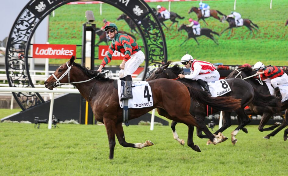 Jockey in red-green silks rides a brown horse with bib 4 ('Tron Bolt') leading a horse race on a green turf track.