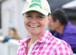 Smiling woman wearing a beige cap with a green logo and a pink checkered shirt, close-up outdoors.
