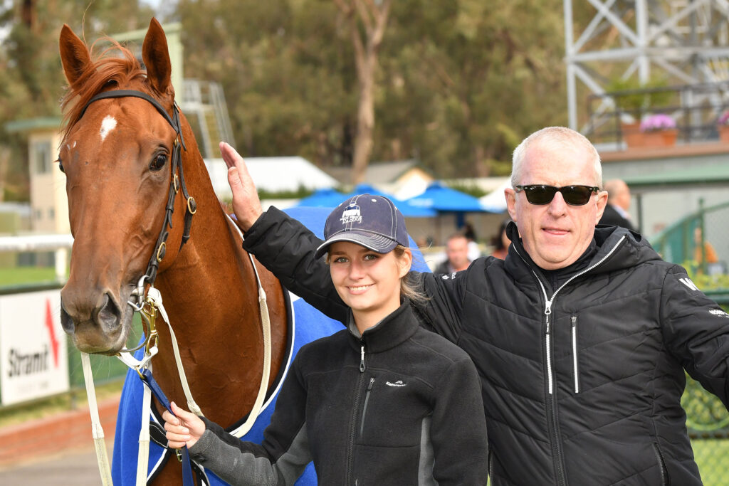 Young woman and an older man pose with a brown horse wearing a blue blanket at an outdoor event.