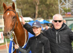 Young woman and an older man pose with a brown horse wearing a blue blanket at an outdoor event.
