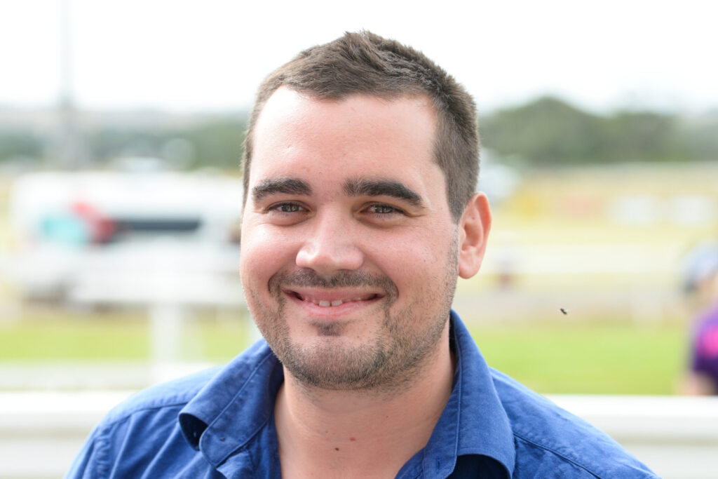 Close-up of a smiling man with short dark hair and stubble wearing a blue shirt outdoors.