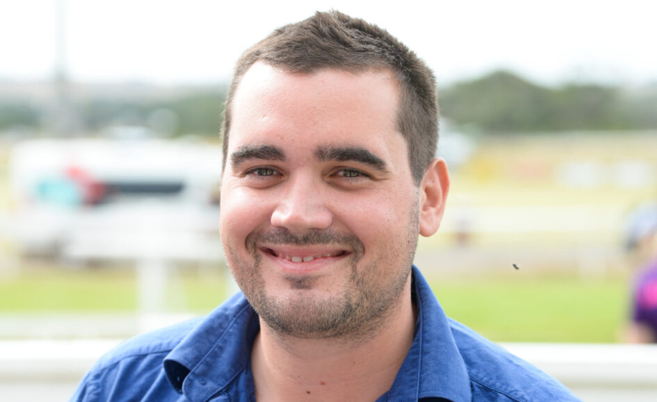 Close-up of a smiling man with short dark hair and stubble wearing a blue shirt outdoors.