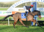 Jockey in a red helmet and black silks rides a horse over a hurdle at a racetrack, with advertising banners in the background.