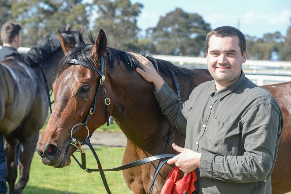 Man smiles while patting a brown horse on a sunny day at the stable area outdoors.