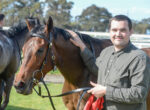 Man smiles while patting a brown horse on a sunny day at the stable area outdoors.