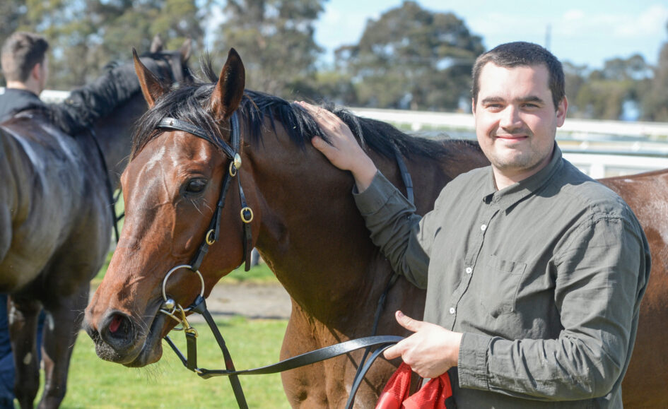 Man smiles while patting a brown horse on a sunny day at the stable area outdoors.