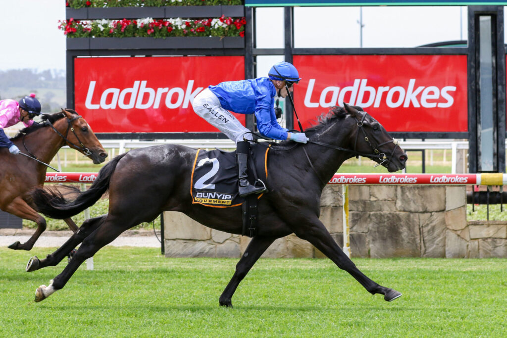 Jockey in blue silks riding a dark horse with number 2, charging down a grass track with red Ladbrokes signs in the background.