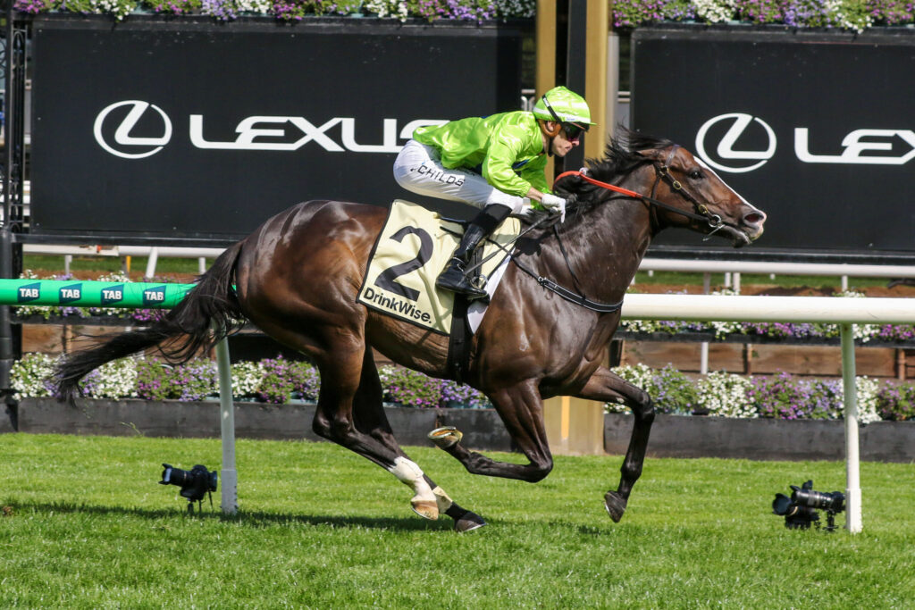 Jockey in neon green silks rides a dark brown horse during a race, with Lexus banners in the background.