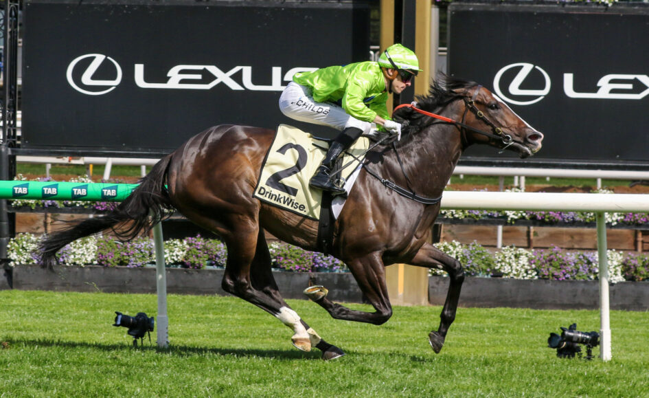 Jockey in neon green silks rides a dark brown horse during a race, with Lexus banners in the background.