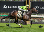 Jockey in neon green silks rides a brown Thoroughbred during a photo finish at a racecourse, Lexus signage in the background.