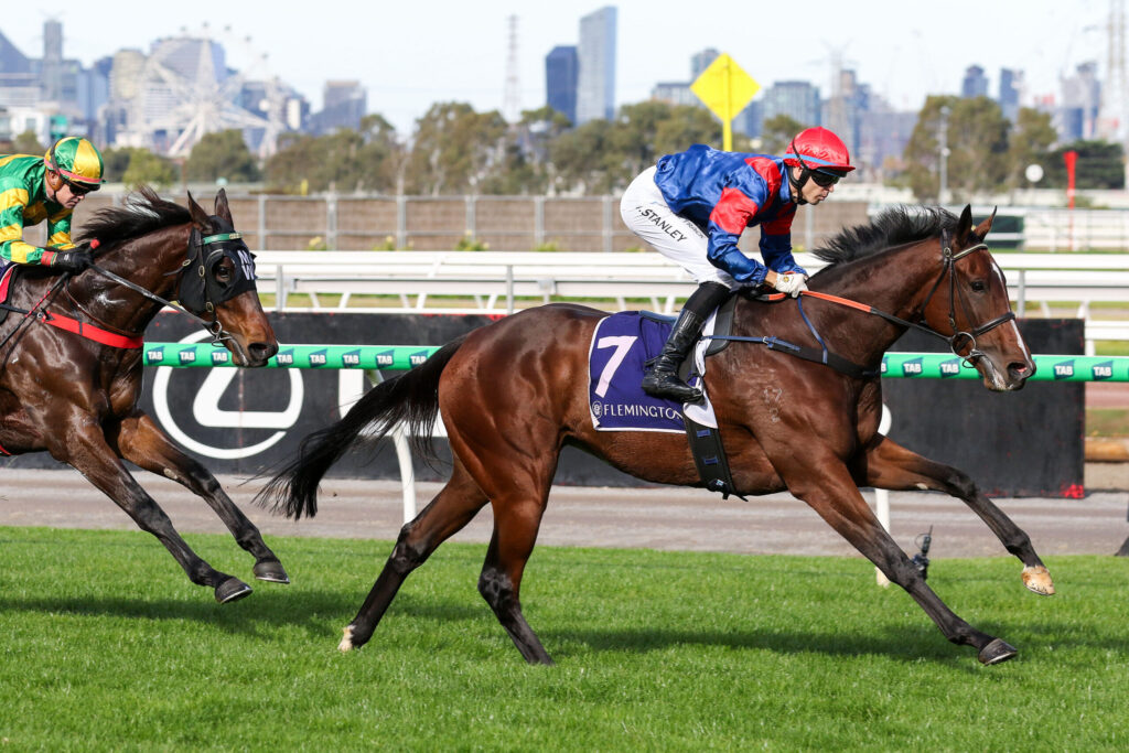 Two jockeys ride racehorses at speed on a green turf track, with a city skyline in the background.