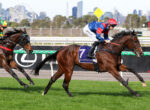 Two jockeys ride racehorses at speed on a green turf track, with a city skyline in the background.