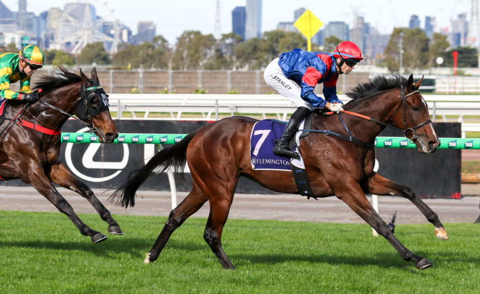 Two jockeys ride racehorses at speed on a green turf track, with a city skyline in the background.