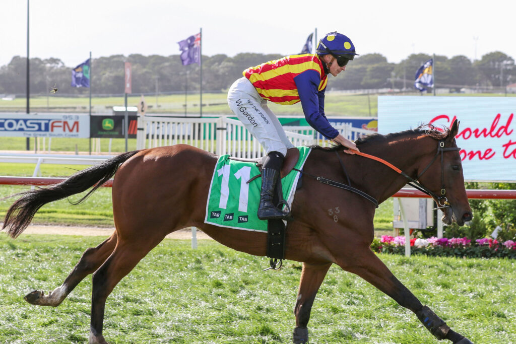 Jockey in red-and-yellow striped silks riding a galloping chestnut horse on a racetrack, green number cloth visible under saddle gearl.