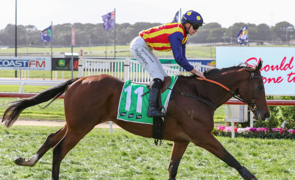 Jockey in red-and-yellow striped silks riding a galloping chestnut horse on a racetrack, green number cloth visible under saddle gearl.