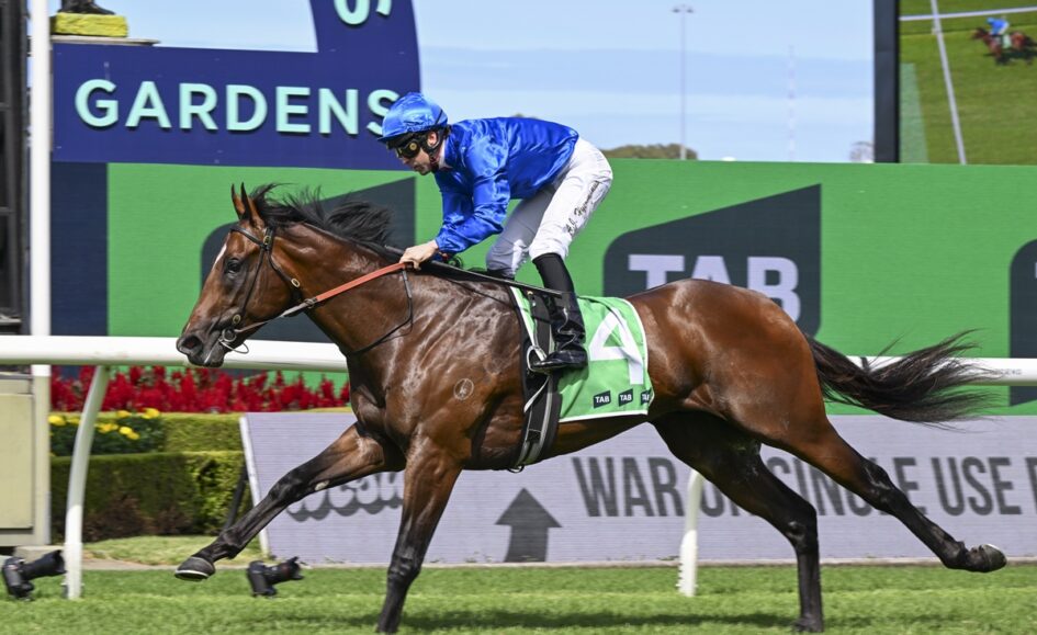 Jockey in blue silks riding a chestnut racehorse with a green number 4 saddlecloth, galloping on a turf track.