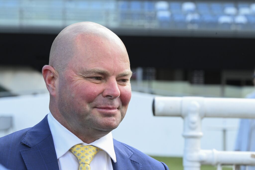 Smiling bald man in a blue suit and yellow patterned tie outdoors, with a white railing behind him.