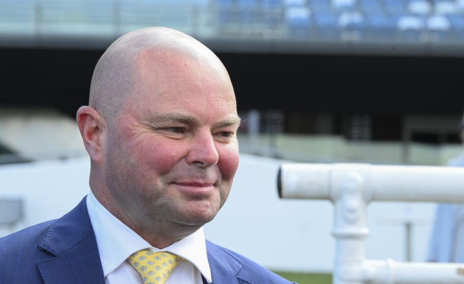 Smiling bald man in a blue suit and yellow patterned tie outdoors, with a white railing behind him.