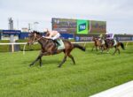 Leading brown racehorse with a peach-sleeved jockey charging on a green turf track; large scoreboard behind. (Kensington signage visible)
